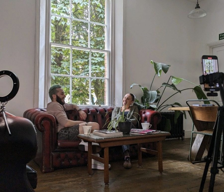 A shot of an office room where two employees sit on a brown leather couch. At the edges of the picture you see a ringlight and a mobile phone that is capturing the moment for EGC content.