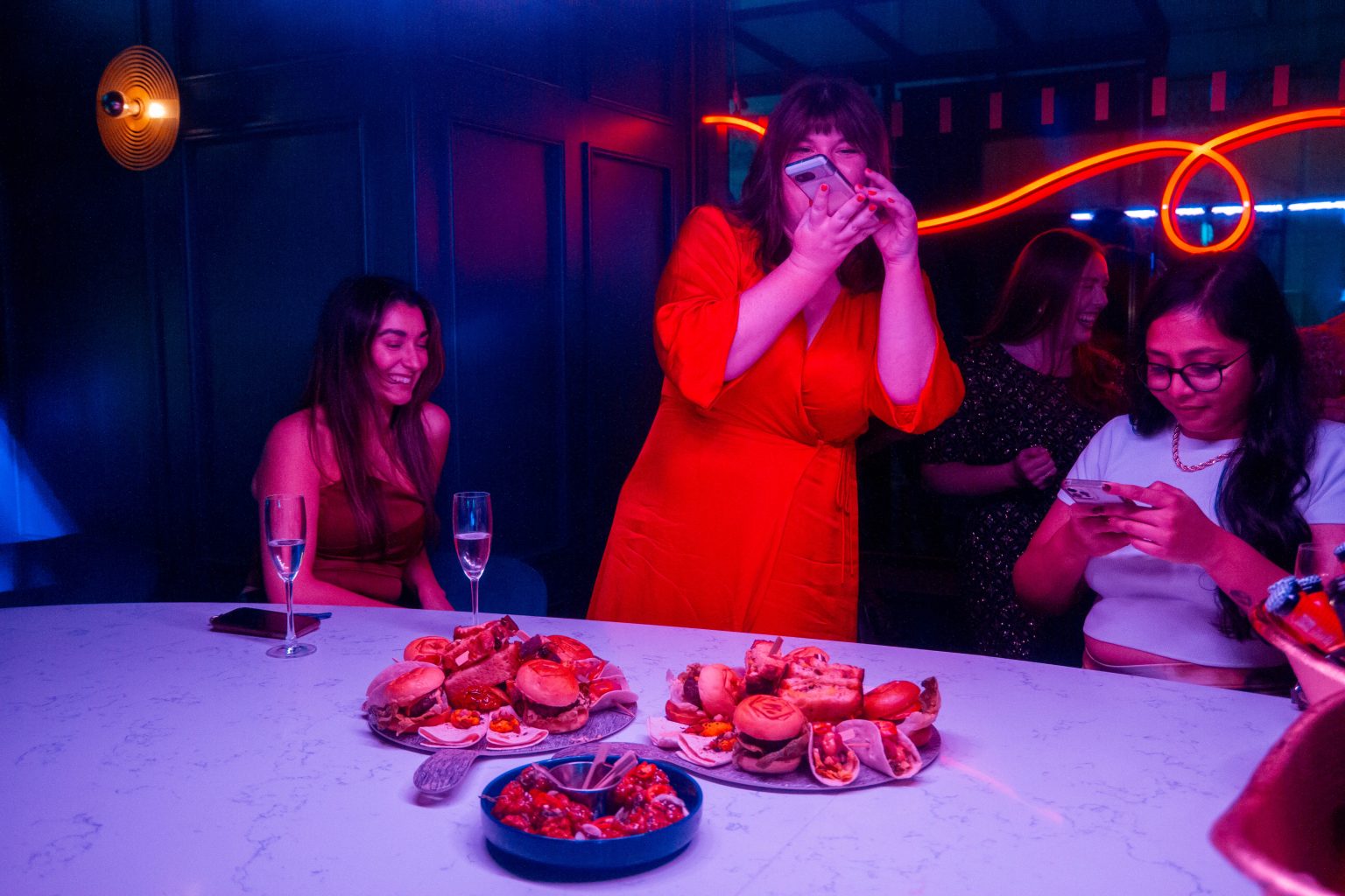 At a bar table, three girls with glasses of complimentary proessco. An influencer wearing a red dress stands up to take a photo of the Manahatta food.