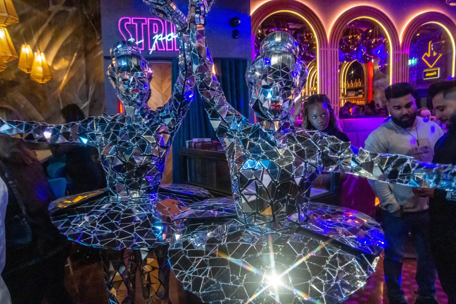 The image gives an impression of the launch party night and shows two party performers in outfits made completely of mirrored geometric shapes, tessellating to fit their bodies.