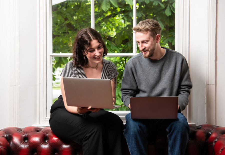 Two employees from marketing agency Tank sit shoulder to shoulder. They sit on the back of a red sofa in front of a window. They both have laptops on their laps and look like they are comparing notes.