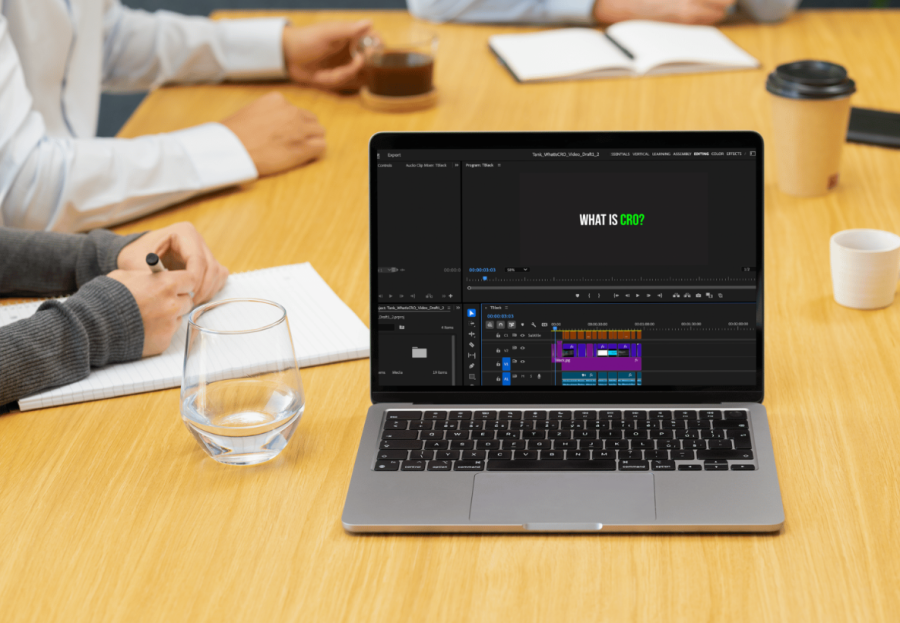 A laptop screen shows video editing software. It is in the foreground on a tabl. The arms of business people in the background suggest a marketing strategy meeting is happening around it.