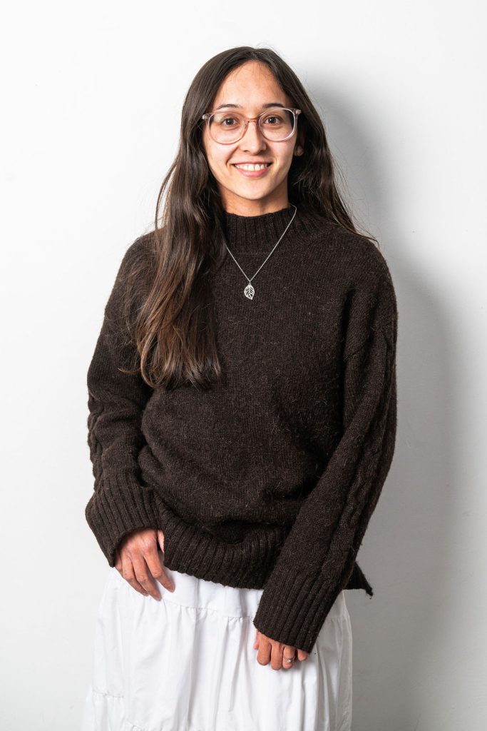 Nadia is a PR executive at Tank. She has long dark brown hair and wears glasses. She is wearing a brown sweater and long white baggy skirt. She is looking into the camera, smiling, with one hand rested on her hip.