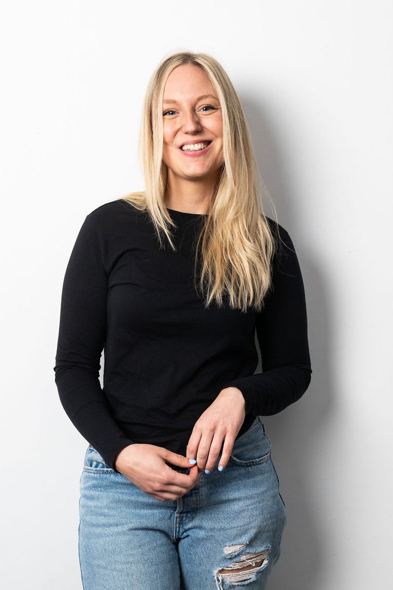 Content marketing manager, Dolly, is a white female with blonde hair. She is standing against a white background, wearing a black top and jeans, smiling into the camera.