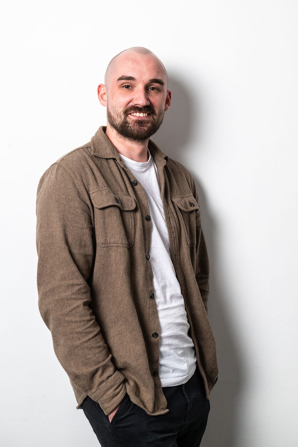 Connor is an SEO manager. He is standing side on with head turned to the camera. He is wearing a brown shirt over white tshirt. He has facial hair and is smiling at the camera.